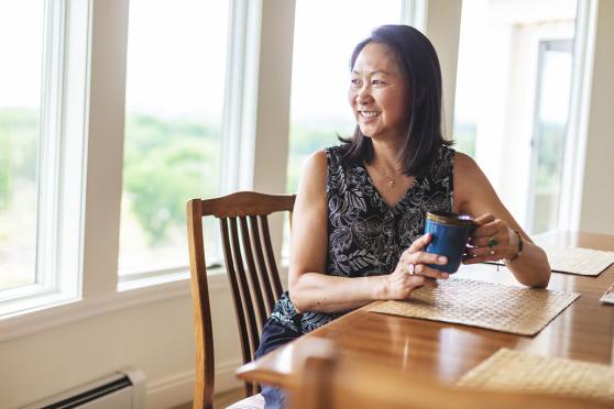 A woman sitting at a table, holding a mug, smiling towards a window